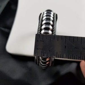Op de afbeelding: Een zilveren armband met een kralenontwerp, gemeten met een liniaal om iets meer dan 2,5 cm breed te zijn.