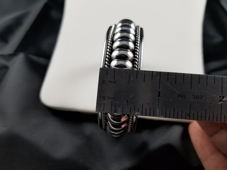 Op de afbeelding: Een zilveren armband met een kralenontwerp, gemeten met een liniaal om iets meer dan 2,5 cm breed te zijn.