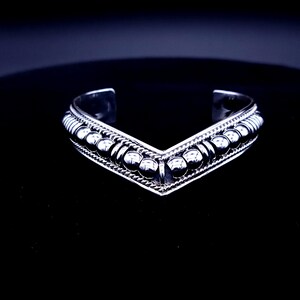 May include: A silver cuff bracelet with a V-shaped design. The bracelet features a row of small, round beads along the top and bottom edges, with a textured pattern in between. The bracelet is displayed on a black surface.