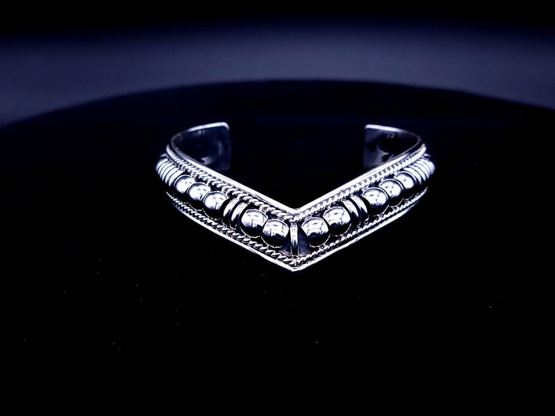 May include: A silver cuff bracelet with a V-shaped design. The bracelet features a row of small, round beads along the top and bottom edges, with a textured pattern in between. The bracelet is displayed on a black surface.