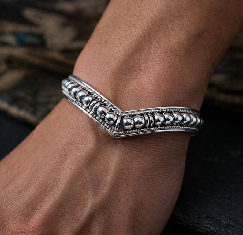 May include: A silver bracelet with a V-shaped design. The bracelet features a series of small, round beads and intricate detailing. The bracelet is worn on a wrist, showcasing its design and craftsmanship.