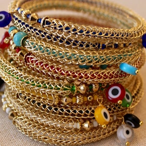 Puede incluir: Una pila de pulseras de alambre dorado con cuentas de colores y amuletos de ojo turco. Las pulseras están superpuestas, creando una exhibición colorida y llamativa.
