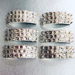 May include: Six silver napkin rings, each decorated with multiple rows of sparkling, clear crystals. The crystals reflect light, creating a dazzling effect. The napkin rings are arranged on a silver surface.
