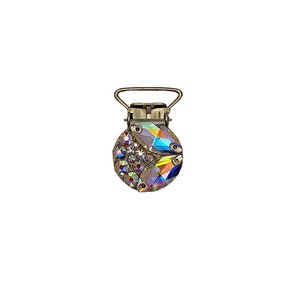 May include: A silver-coloured pacifier clip with a circular, iridescent crystal design. The clip features a metal clasp and a loop for attaching to a soother or other item. The crystals reflect a rainbow of colours.