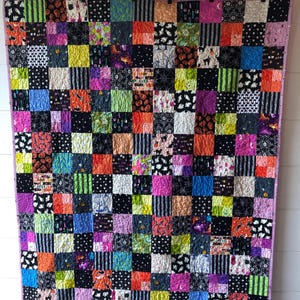 May include: A colorful patchwork quilt with a variety of Halloween-themed fabrics. The quilt features squares in black, orange, purple, green, and pink, with patterns including stripes, spiderwebs, and bats. The quilt is hung on a wall.