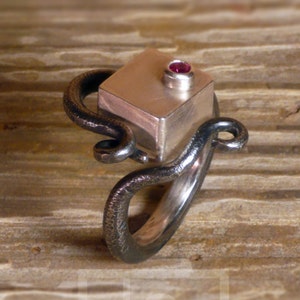 Peut inclure: Une bague en argent avec une pierre de forme carrée sertie sur le dessus. La pierre est de couleur rose foncé. La bande de la bague est en métal torsadé.