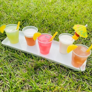 May include: Five colourful cocktails in clear plastic cups with straws and garnishes on a white tray. The tray is on a bed of green grass.
