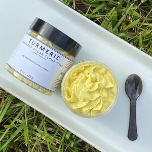 Puede incluir: Un plato de cerámica blanco con un exfoliante de azúcar batido amarillo en un frasco de vidrio. El frasco tiene una tapa negra y una etiqueta blanca que dice "Turmeric Whipped Sugar Scrub Soap" con el texto "Cleanse | Exfoliate" y "Rejuvenating | Uplifting + Herbal" debajo. El frasco está etiquetado con "4 oz | 113 g". Una cuchara negra está en el plato.