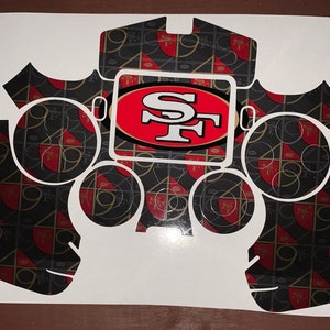 May include: A white sticker sheet with black and red patterned designs for a video game controller. The sticker sheet features the San Francisco 49ers logo in red and white.
