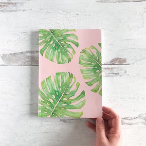 May include: A pink notebook with a green monstera leaf pattern. The notebook is closed and has a blank cover.
