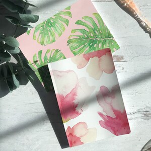 May include: Two notebooks with different floral designs. One notebook has a pink background with green monstera leaves. The other notebook has a white background with pink watercolor flowers.