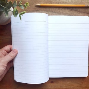 May include: An open lined notebook with white pages and a white cover. The notebook is lying open on a wooden surface.