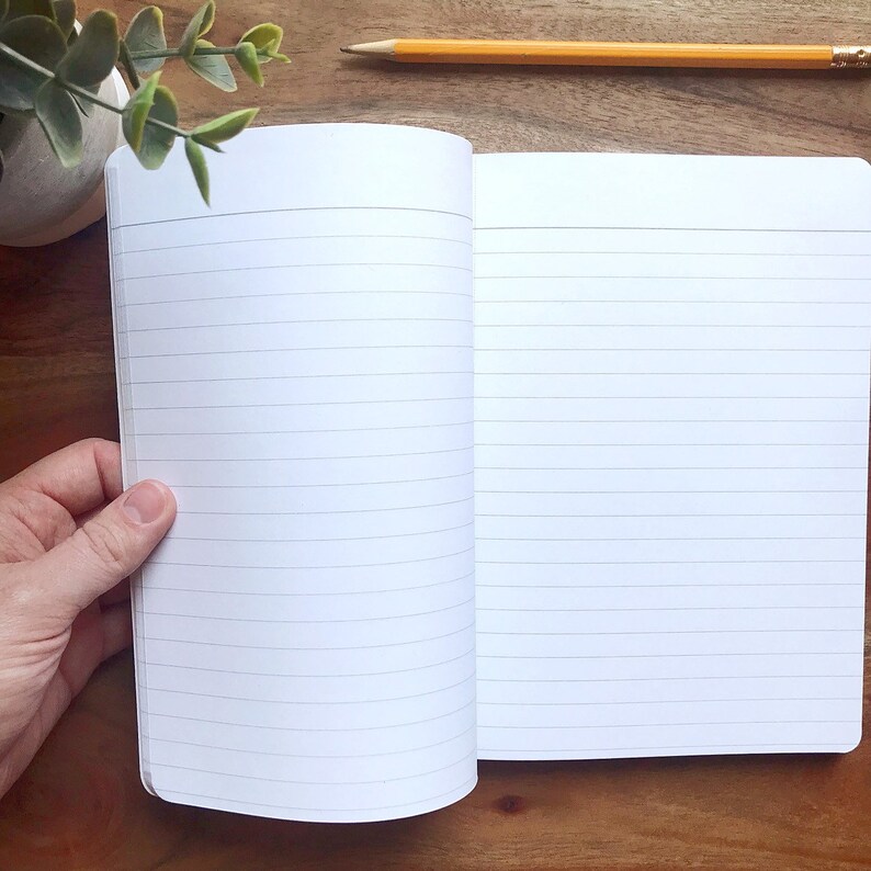 May include: An open lined notebook with white pages and a white cover. The notebook is lying open on a wooden surface.