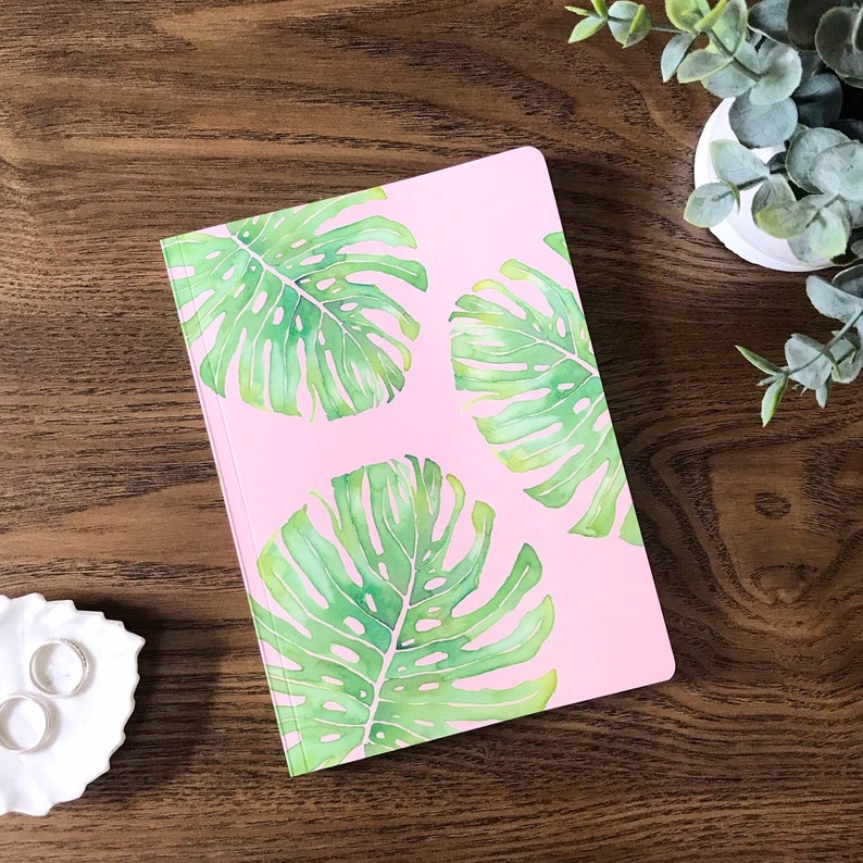 May include: A pink notebook with a tropical leaf design. The leaves are green and have a watercolor effect.