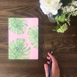 May include: A pink notebook with a tropical leaf design. The notebook is on a wooden surface with a pen and a bouquet of white flowers.
