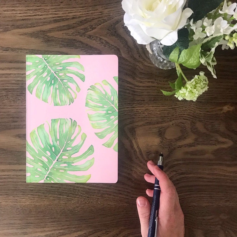 May include: A pink notebook with a tropical leaf design. The notebook is on a wooden surface with a pen and a bouquet of white flowers.