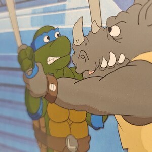 May include: A colorful animation cel featuring a green turtle character with a blue mask and belt holding a sword, engaged in a confrontation with a grey rhinoceros character. The scene is set against a blue background with geometric shapes.
