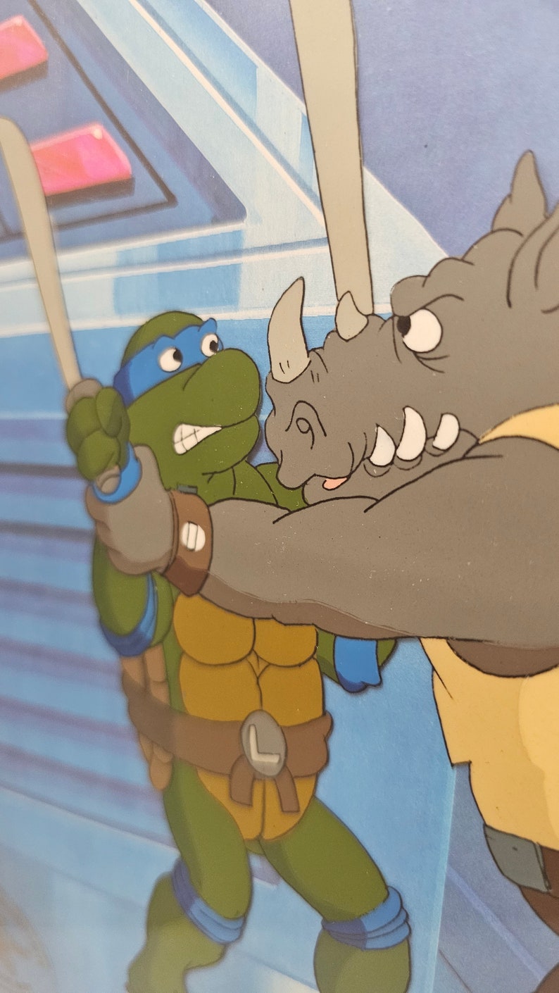 May include: A colorful animation cel featuring a green turtle character with a blue mask and belt holding a sword, engaged in a confrontation with a grey rhinoceros character. The scene is set against a blue background with geometric shapes.