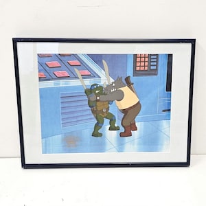 May include: A framed animation cel featuring a green turtle character with a blue mask and a gray rhinoceros character in a tan shirt and brown pants. The two characters are engaged in a sword fight inside a blue room with a control panel.