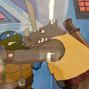 May include: A vintage animation cel featuring a green turtle character with a blue mask and sword engaged in a confrontation with a grey rhinoceros character wearing a yellow shirt. The background shows a blue control panel with red buttons.