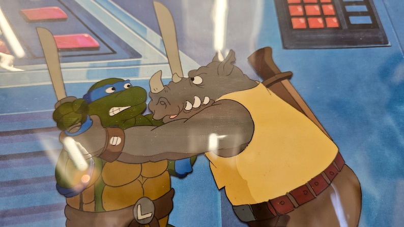 May include: A vintage animation cel featuring a green turtle character with a blue mask and sword engaged in a confrontation with a grey rhinoceros character wearing a yellow shirt. The background shows a blue control panel with red buttons.