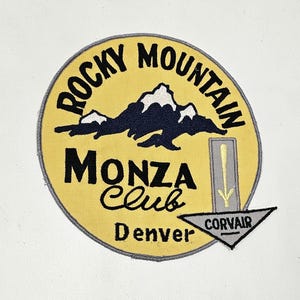 May include: A circular embroidered patch with a yellow background and a grey border. It features a black mountain graphic with white snow caps, the text "ROCKY MOUNTAIN MONZA Club Denver" in black, and a grey arrow pointing down with the word "CORVAIR".