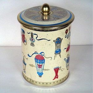 May include: A vintage cream-colored metal canister with a gold lid and a whimsical design featuring hot air balloons, a chair, a boot, and a carriage. The canister has a gold trim and a star pattern around the bottom.