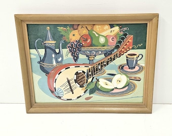 May include: A framed painting depicting a still life with a mandolin, fruit bowl, tea set, and sliced apples. The mandolin is red and cream-colored. The fruit bowl contains grapes, pears, and other fruits. The tea set is blue and gold.