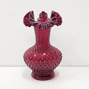 May include: A red glass vase with a ruffled top and a textured, dotted surface. The vase is a classic design with a vintage look.