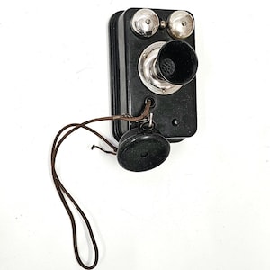 May include: A black vintage wall-mounted phone with a black handset and a cord. The phone has two silver bells and the text "The Couch Co.  Boston" on the front.