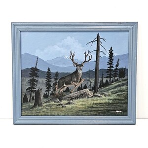 May include: A framed painting of a deer leaping over a log in a forest scene. The artwork features a buck with large antlers, set against a backdrop of mountains, trees, and a blue sky. The frame is a light blue color.