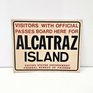May include: A vintage sign with red and black text on a cream background. The sign reads "VISITORS WITH OFFICIAL PASSES BOARD HERE FOR ALCATRAZ ISLAND UNITED STATES GOVERNMENT FEDERAL BUREAU OF PRISONS".