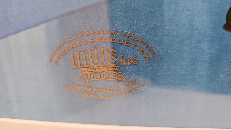 May include: A blue animation art cel featuring a gold embossed seal that reads ORIGINAL PRODUCTION MWS inc TEENAGE MUTANT NINJA TURTLES ANIMATION ART, with additional text including 1985 MIRAGE STUDIOS and 1988-89 MURAKAMI WOLF SWENSON INC.