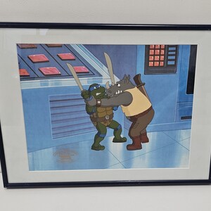 May include: A framed animation cel featuring a blue-masked turtle character holding a sword and grappling with a grey rhinoceros character in a tan vest. The scene is set against a blue background with a control panel and a gold seal in the corner.