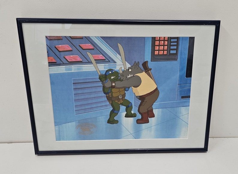May include: A framed animation cel featuring a blue-masked turtle character holding a sword and grappling with a grey rhinoceros character in a tan vest. The scene is set against a blue background with a control panel and a gold seal in the corner.