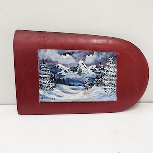 May include: A red, oval-shaped piece of metal with a painted winter scene of snow-capped mountains and trees. The painting is signed "Strandlf" in the bottom left corner.