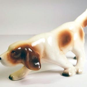 May include: A white porcelain figurine of a brown and white dog with brown spots. The dog is in a running pose with its head turned to the side.