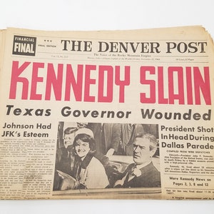 May include: A black and white newspaper headline announcing the assassination of President John F. Kennedy. The headline reads "Kennedy Slain" in large red letters. The sub-headline reads "Texas Governor Wounded".