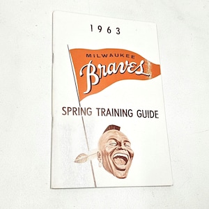 May include: A 1963 Milwaukee Braves Spring Training Guide booklet with a white cover. The cover features an orange pennant flag graphic with the text "Milwaukee Braves" in white script, and a brown illustration of a smiling person with a feather.
