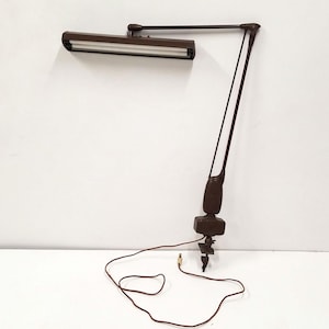 May include: A brown adjustable desk lamp with a rectangular light fixture. The lamp has a clamp base and a flexible arm. The cord is brown. The lamp is designed for task lighting.