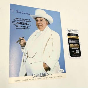 May include: A glossy photograph of Sorrell Booke as Boss Hogg from The Dukes of Hazzard, wearing a white suit and hat, holding a cigar. The print features a handwritten inscription to Bob Inman and a signature, accompanied by a JSA certification card.
