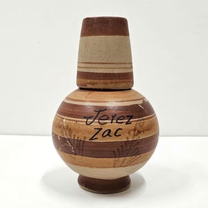 May include: A brown and tan ceramic bottle with a lid. The bottle has a striped pattern and the words "Jerez Zac" written on it.