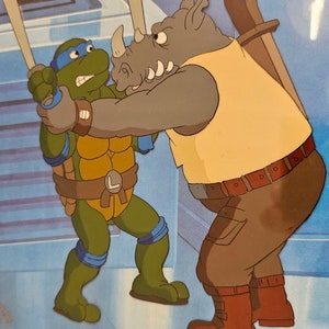 May include: A vintage animation cel featuring a green turtle character with a blue mask and belt with an L emblem, grappling with a large grey rhinoceros character wearing a yellow tank top and brown pants against a blue background.