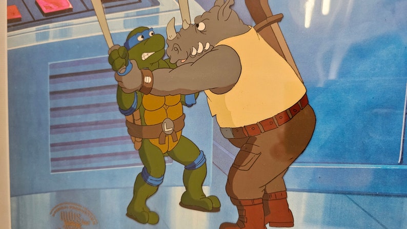 May include: A vintage animation cel featuring a green turtle character with a blue mask and belt with an L emblem, grappling with a large grey rhinoceros character wearing a yellow tank top and brown pants against a blue background.