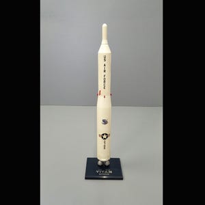 May include: A white model of a Titan rocket with the words "US AIR FORCE" and "TITAN" printed on the side. The rocket is on a blue base.