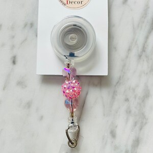 May include: A pink and iridescent beaded retractable badge holder with a clear plastic badge clip and a silver metal swivel clip. The holder is attached to a white card with the text "Lyon Street Studio ID Badge Decor".
