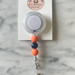 May include: A clear retractable badge holder with a white fabric insert. The holder is attached to a black and orange beaded lanyard with a silver clip.