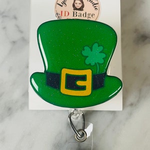 Op de afbeelding: Groene glitter-leprechaunhoed met een gouden gesp en een klavertje vier op de bovenkant. De hoed is bevestigd aan een transparante, intrekbare badgehouder van plastic met een zilveren clip.
