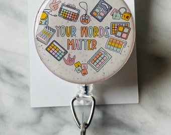 May include: A white retractable badge holder with a round, colorful button featuring the text "YOUR WORDS MATTER" surrounded by various colorful illustrations of musical equipment and a peace sign.