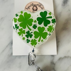 Op de afbeelding: Een witte hartvormige badgehouder met een groen klaverbladpatroon en glitter. De badgehouder is bevestigd aan een transparante intrekbare badgehaspel.